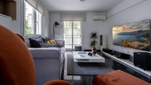 A modern living room with a sofa, coffee table, and a large TV on a white wall. - The Black Marble - Sea View / 2BR / 1,5BA (Paleo Faliro)