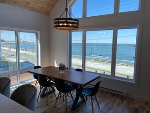 Un comedor con mesa y sillas de madera, ventanas amplias con vista a la playa y al océano, y una lámpara colgante.