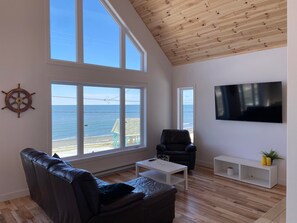 Un salón amplio con un gran ventanal con vistas al mar, un sofá de cuero negro, una mesa de centro blanca y un televisor montado en la pared.