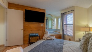 Un dormitorio con cama, pared de madera, televisor montado arriba y baño con bañera, además de una ventana con cortinas.