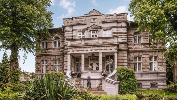 Ein großes, historisches Gebäude mit einem prunkvollen Eingang, zahlreichen Fenstern und einer zentralen Treppe, die zu einem Balkon führt.