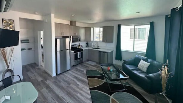 A modern kitchen with stainless steel appliances, a dining area with a glass table, and a living room with a dark green sofa. - One Bedroom Hideaway (Reno)