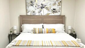 Una cama bien hecha con ropa blanca y almohadillas decorativas, un cabecero de madera y una obra de arte floral sobre ella.