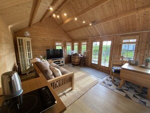 A cozy cabin interior with a wooden ceiling, a TV, a sofa, a dining table, and a large window offering a view of the outdoors. - Character Detached 3-bed Cottage Audlem Cheshire (Crewe)