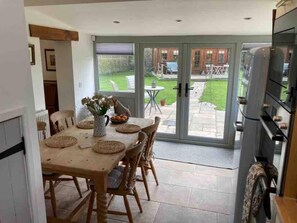 A dining area with a table set for four, a vase with dried flowers, and a view of a garden through glass doors.