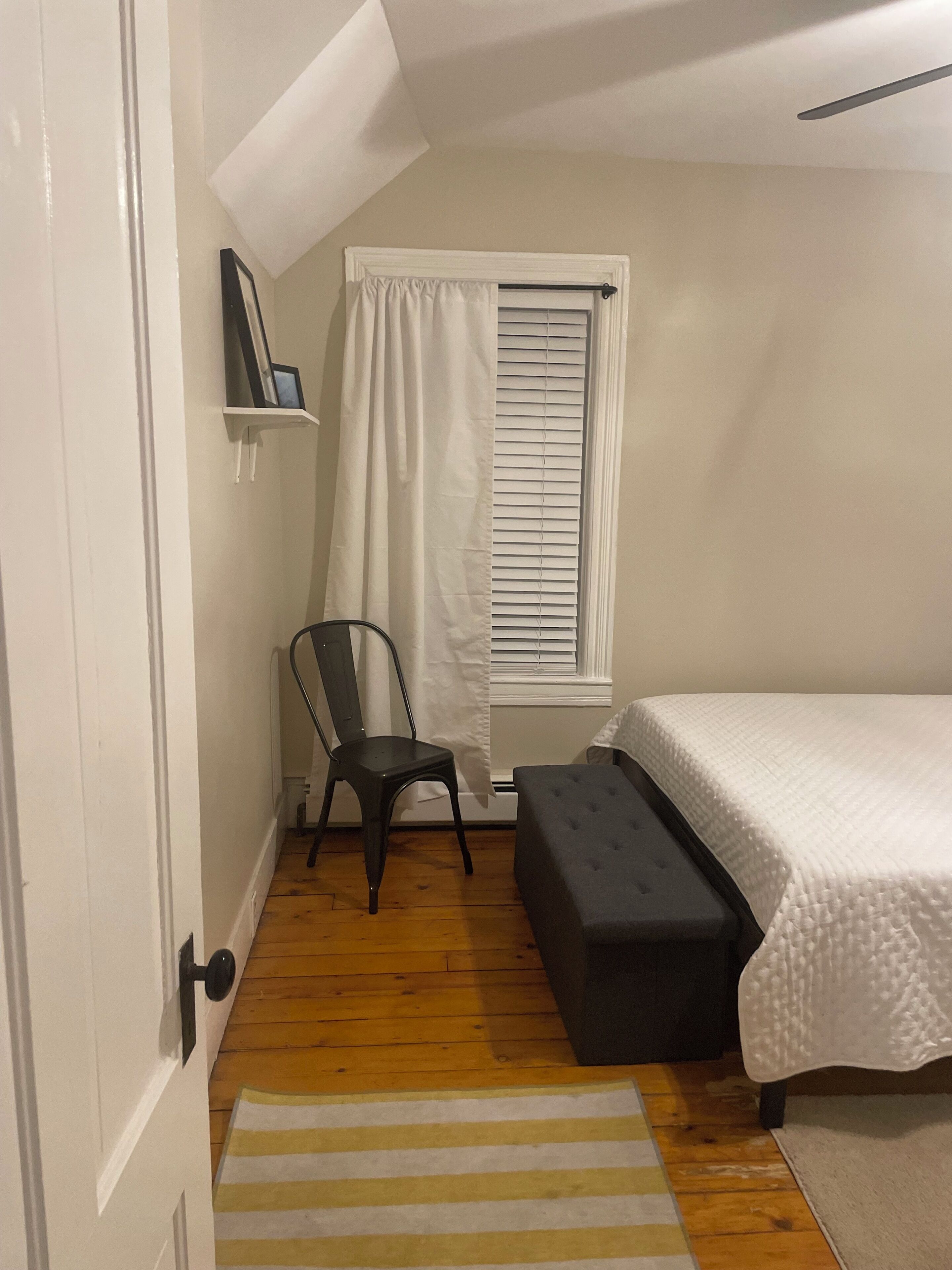 Une chambre à coucher avec un lit, une chaise, une fenêtre avec des stores, une étagère avec un cadre photo et un tapis à rayures.