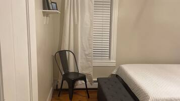 Une chambre à coucher avec un lit, une chaise, une fenêtre avec des stores, une étagère avec un cadre photo et un tapis à rayures.