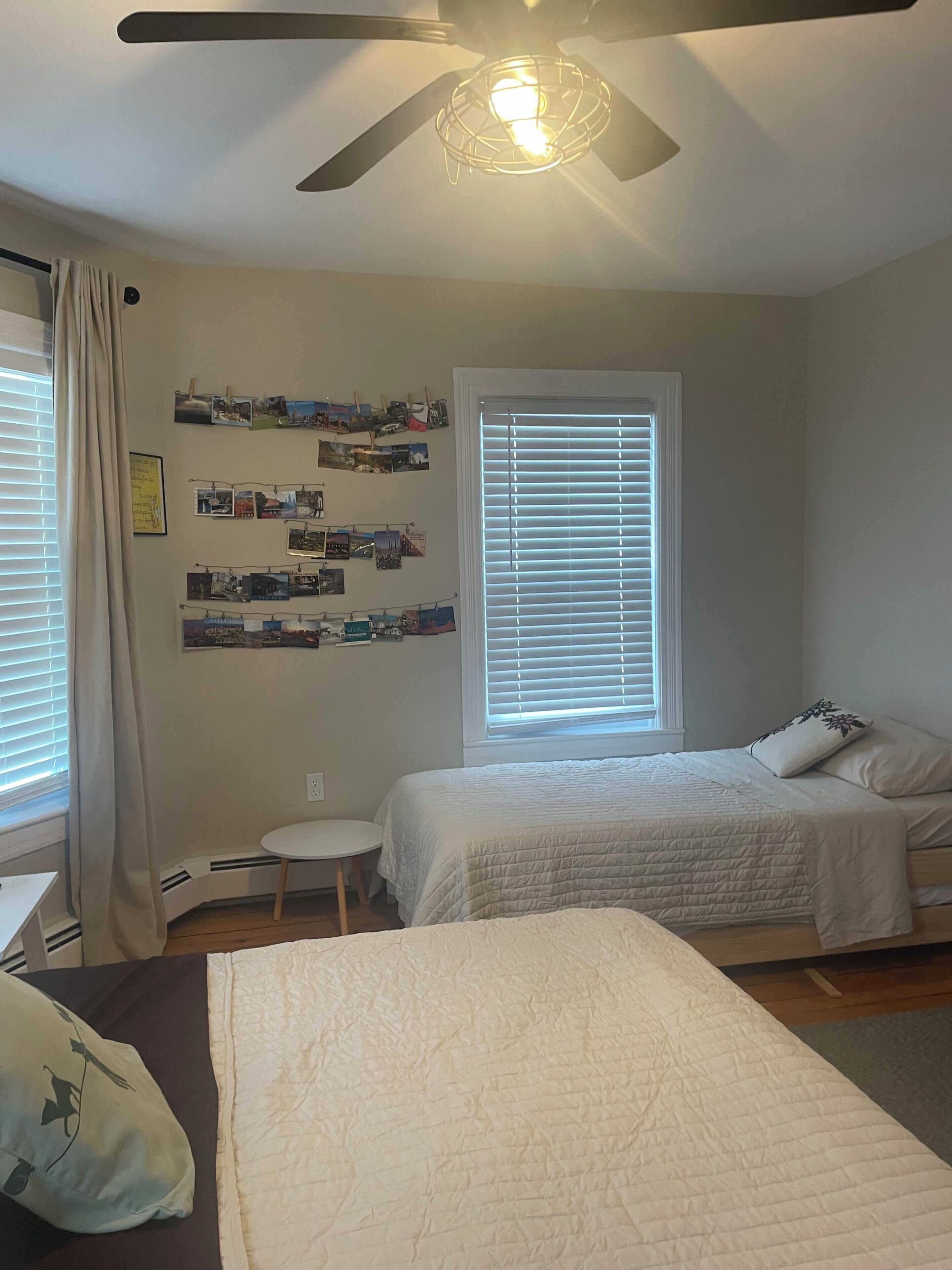 A bedroom with two beds, a ceiling fan, a wall-mounted light fixture, and a window with blinds.