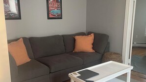 Un salon avec un canapé gris, deux coussins orange, une table basse blanche et deux affiches encadrées au mur.