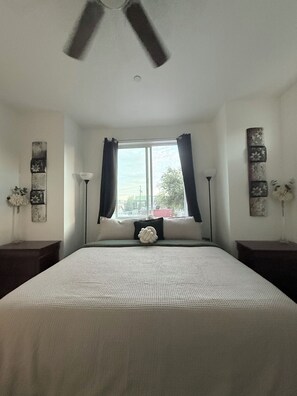 A bedroom with a bed, a window with curtains, a ceiling fan, and wall art.