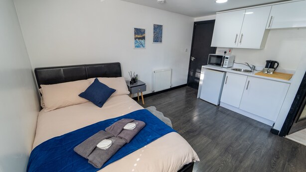 Ett modernt hotellrum med en säng, ett litet sängbord, en mikrovågsugn, en vattenkokare och ett litet pentry.