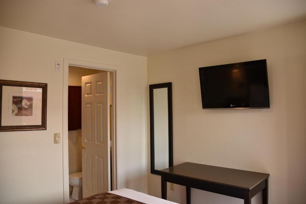 A hotel room with a bed, a television mounted on the wall, a door leading to a bathroom, and a framed picture on the wall.