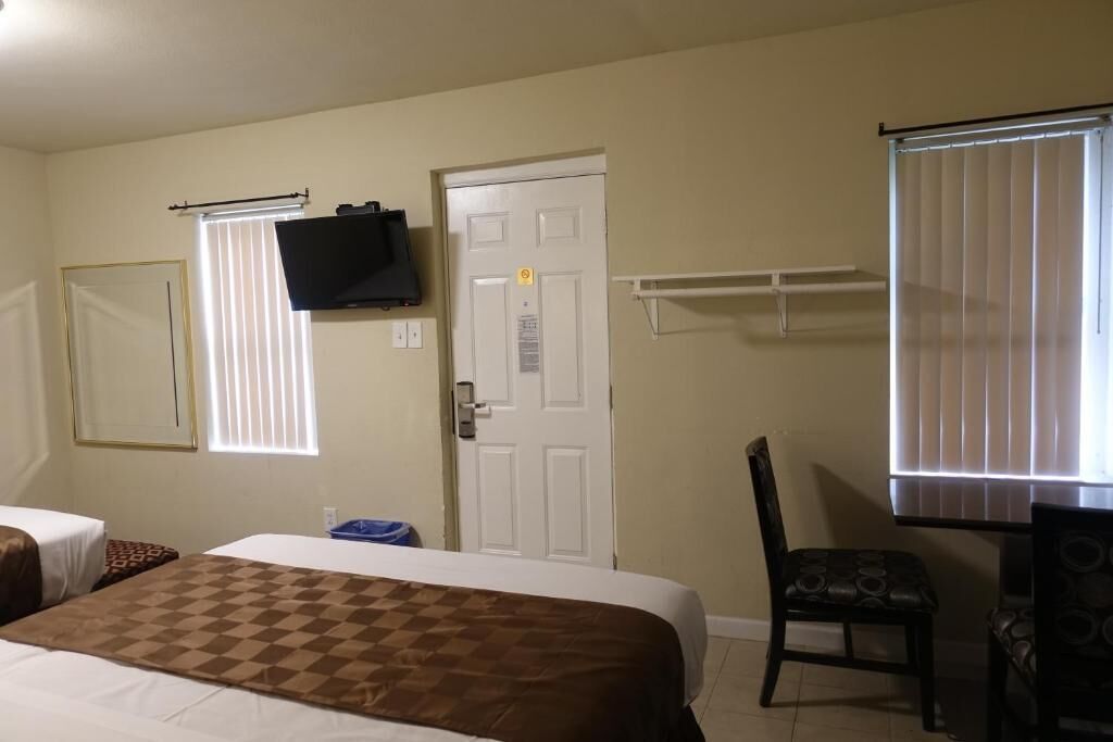 A hotel room with a bed, a television mounted on the wall, a door with a lock, a window with blinds, and a chair.