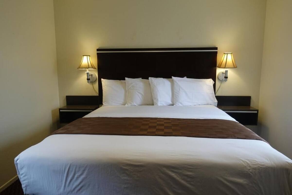 A neatly made bed with white linens and a brown accent, flanked by two bedside tables with lamps, in a simple room with plain walls.