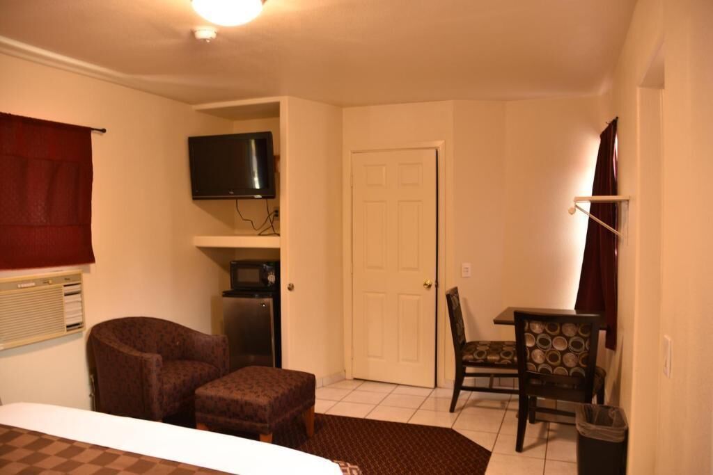 A small, well-lit room with a bed, a chair, a table, a TV, and a door.