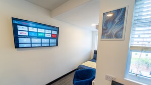 A hotel room with a mounted TV displaying a home screen, a bed with a blue blanket, a window with blinds, and a wall-mounted artwork. - Modern Studio 10 Mins on Train to City! (Birmingham)