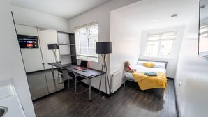 A compact hotel room with a bed, a desk, and a chair. There is a window with blinds, a lamp, and a teddy bear on the bed. - Stunning Studio Next to Restaurants! (Birmingham)