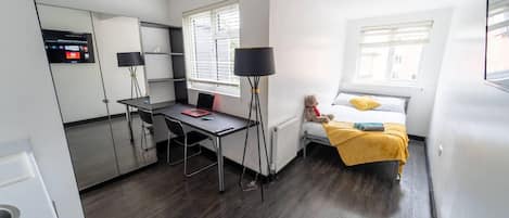 A compact hotel room with a bed, a desk, and a chair. There is a window with blinds, a lamp, and a teddy bear on the bed.