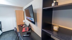 Ein modernes Hotelzimmer mit einem flachbildfernseher an der Wand, einem Schreibtisch mit einem Laptop, einer Fernbedienung und einem kleinen dekorativen Gegenstand auf dem Regal.