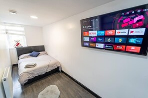 A modern hotel room with a large digital display on the wall, a neatly made bed, a small rug, and a window with a teddy bear.