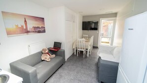 Un espace de vie compact comprenant un canapé gris, une table à manger avec des chaises, un petit coin cuisine et une grande fenêtre.