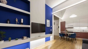Een moderne woonkamer met een televisie, blauwe kasten, een eethoek met stoelen en een keuken op de achtergrond.