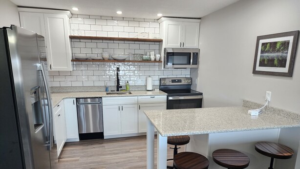 A modern kitchen with white cabinets, a stainless steel refrigerator, a microwave, and a dishwasher. There are wooden stools at the island and a framed picture on the wall. - Green Lake Condo in the heart of Spicer (Spicer)