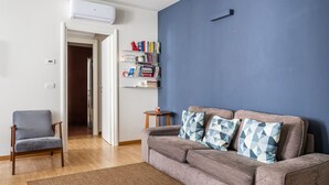 Ein Wohnzimmer mit grauem Sofa, einem kleinen Sessel, einem Bücherregal und einer blauen Wand.