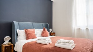 Ein ordentlich bezogenes Bett mit blauem Kopfteil, weißen und rosafarbenen Kissen und gefalteten weißen Handtüchern auf einer roten Bettdecke.