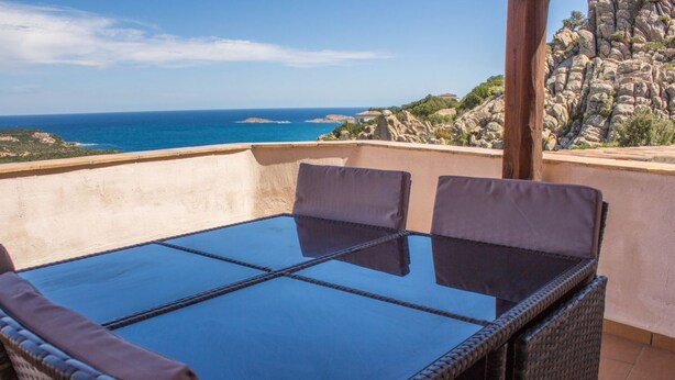 Un balcon doté d’une table à plateau en verre et de chaises en osier, offrant une vue sur une côte rocheuse et la mer.