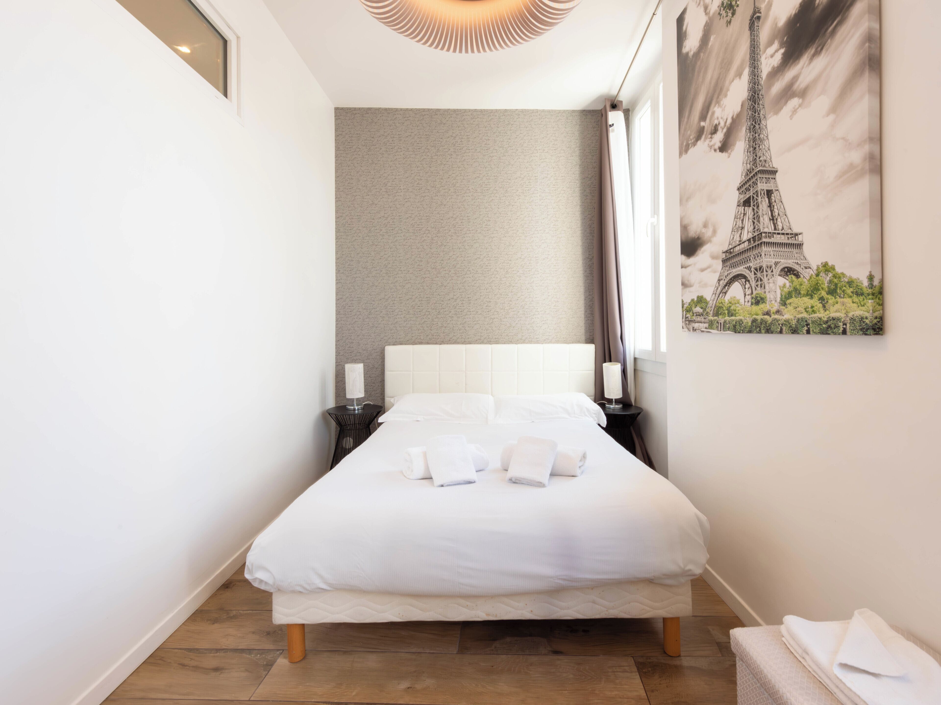 A neatly made bed with white linens and pillows, a bedside lamp, and a framed picture of the Eiffel Tower on the wall.
