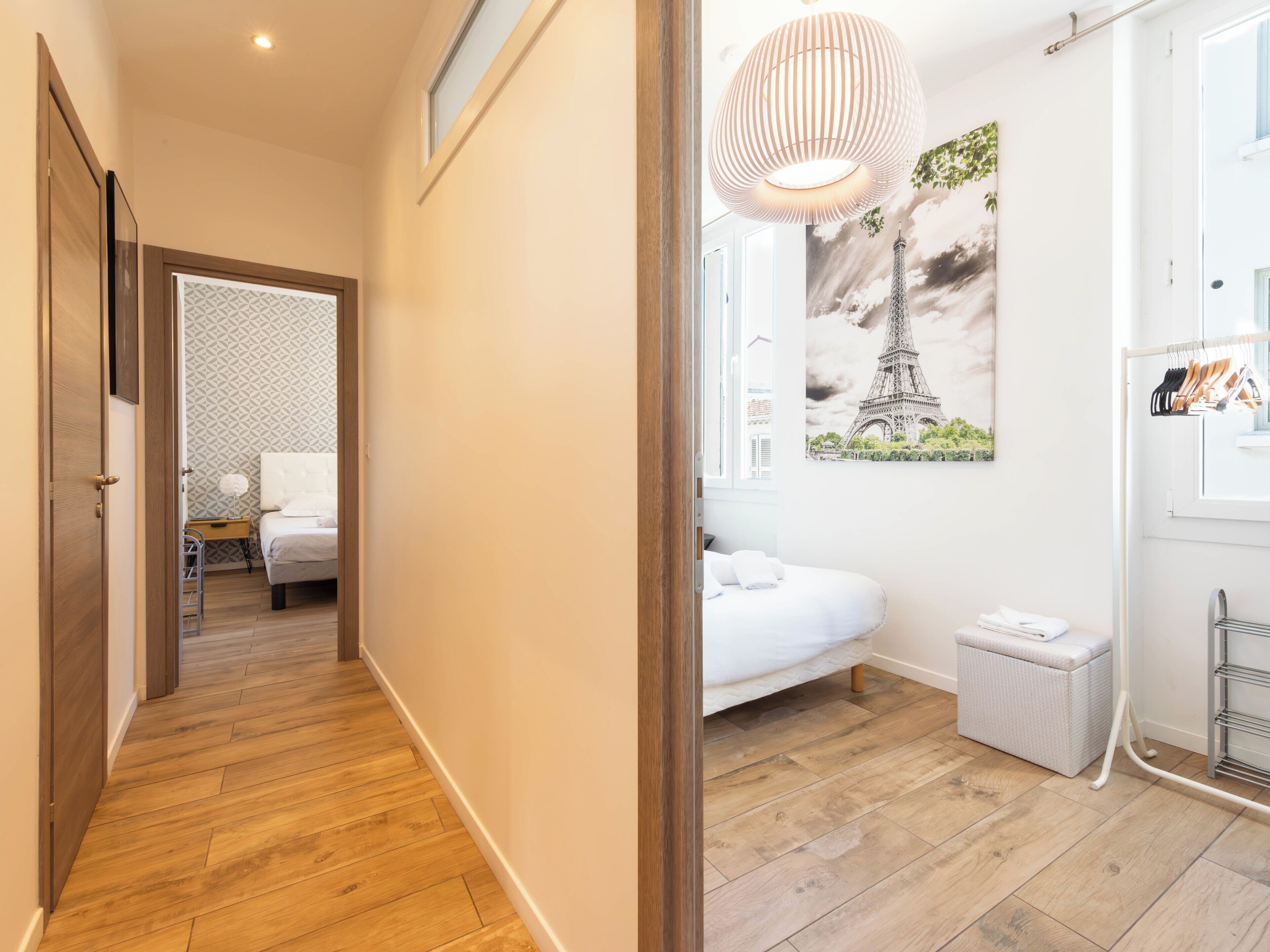 A hotel room with a wooden floor, a bed with white bedding, a small bedside table with a lamp, a wall-mounted radiator, and a window with a view of the Eiffel Tower.