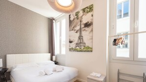 A neatly made bed with white linens, a wall-mounted artwork of the Eiffel Tower, a small bedside table with a lamp, and a window with a view of the outside. - Downtown Cannes, walk to Beaches and Croisette (Cannes)