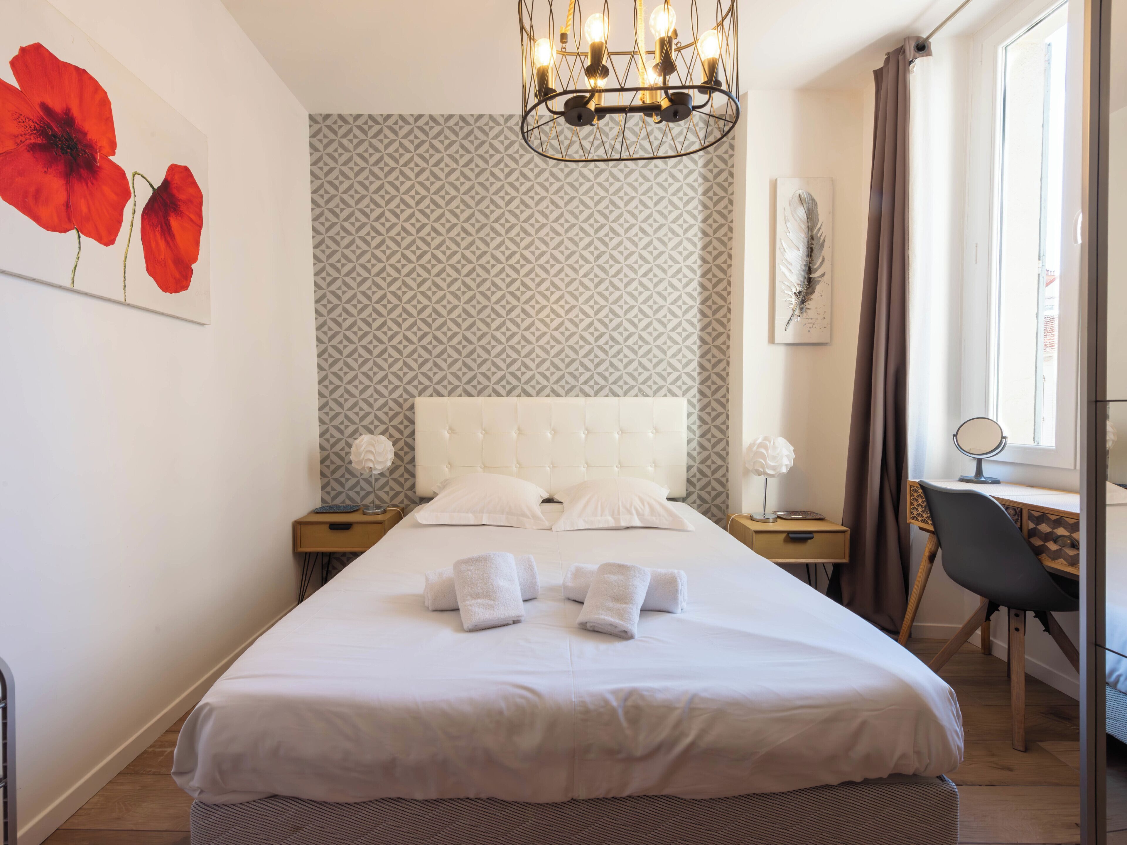A neatly made bed with white linens and towels, a tufted headboard, a geometric patterned wall, a modern chandelier, a small bedside table with a lamp, and a window with curtains.