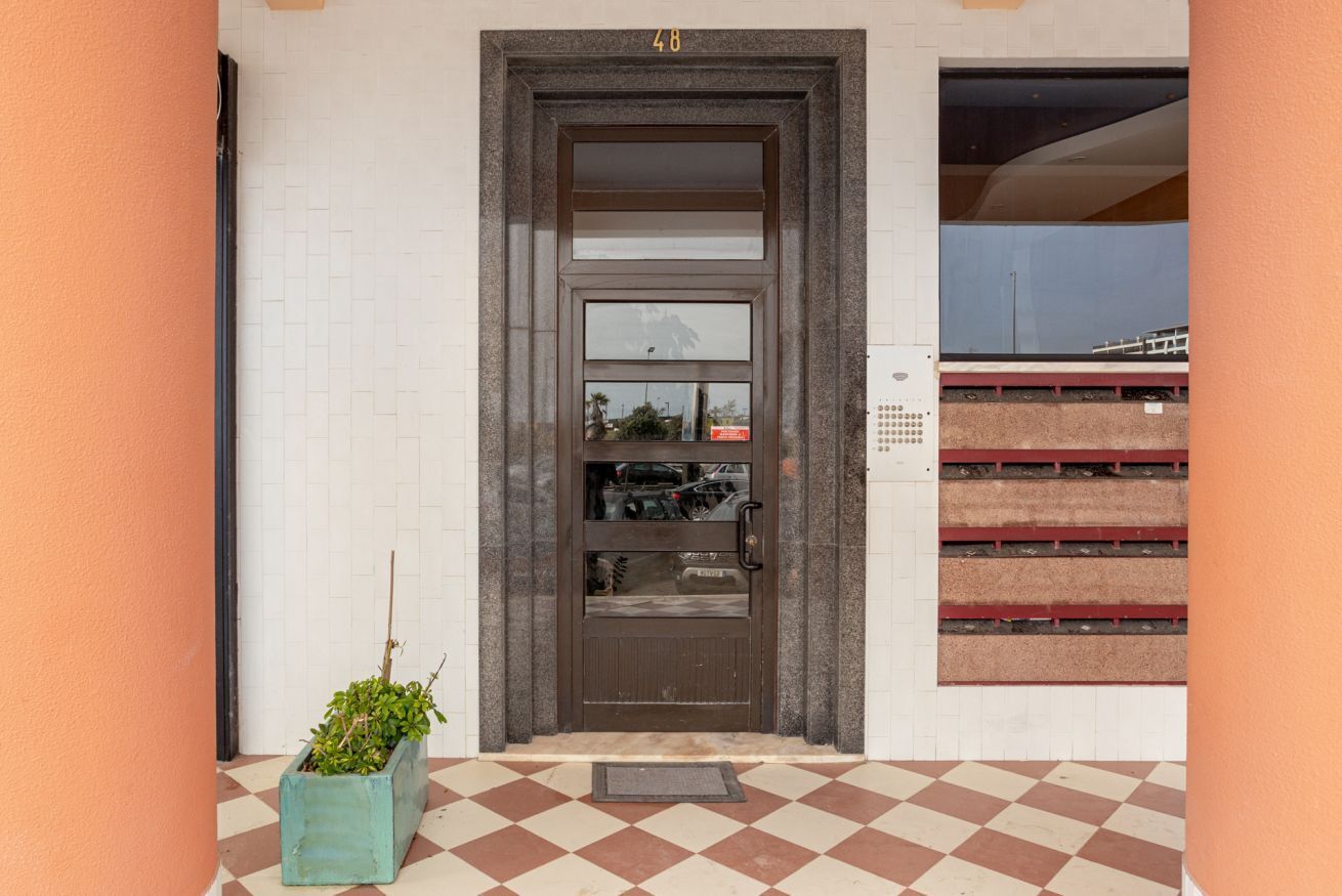 Entrada de um prédio com piso xadrez, uma porta com placa de número e uma pequena planta ao lado.