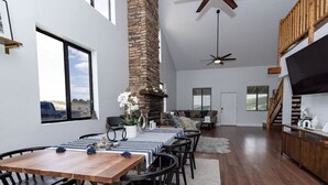 Amplia sala de estar con techos altos, chimenea de piedra, mesa de comedor con sillas, sofá y televisor montado en la pared.