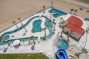 An aerial view of a resort with multiple swimming pools, palm trees, and a red-roofed building.