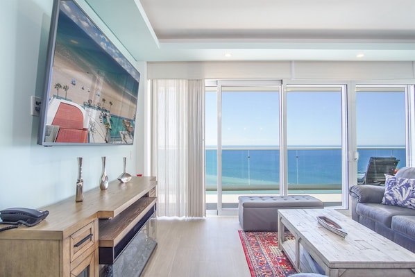 A modern hotel room with a large balcony, a wooden desk, a sofa, and a large painting of a beach scene.