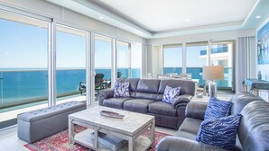 A spacious balcony with a view of the ocean, a grey leather sofa set, a wooden coffee table, and a patterned rug.