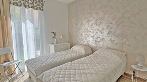 A bedroom with two beds, a nightstand, a lamp, and a window with curtains. - Apartment for 6 people - Air conditioning - Wifi - Downtown - Sainte Maxime (STE MAXIME)