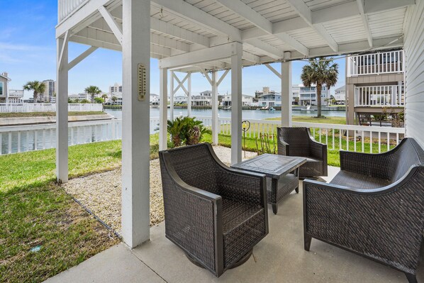 A covered outdoor seating area with wicker furniture, a table, and a view of a body of water and buildings in the background.