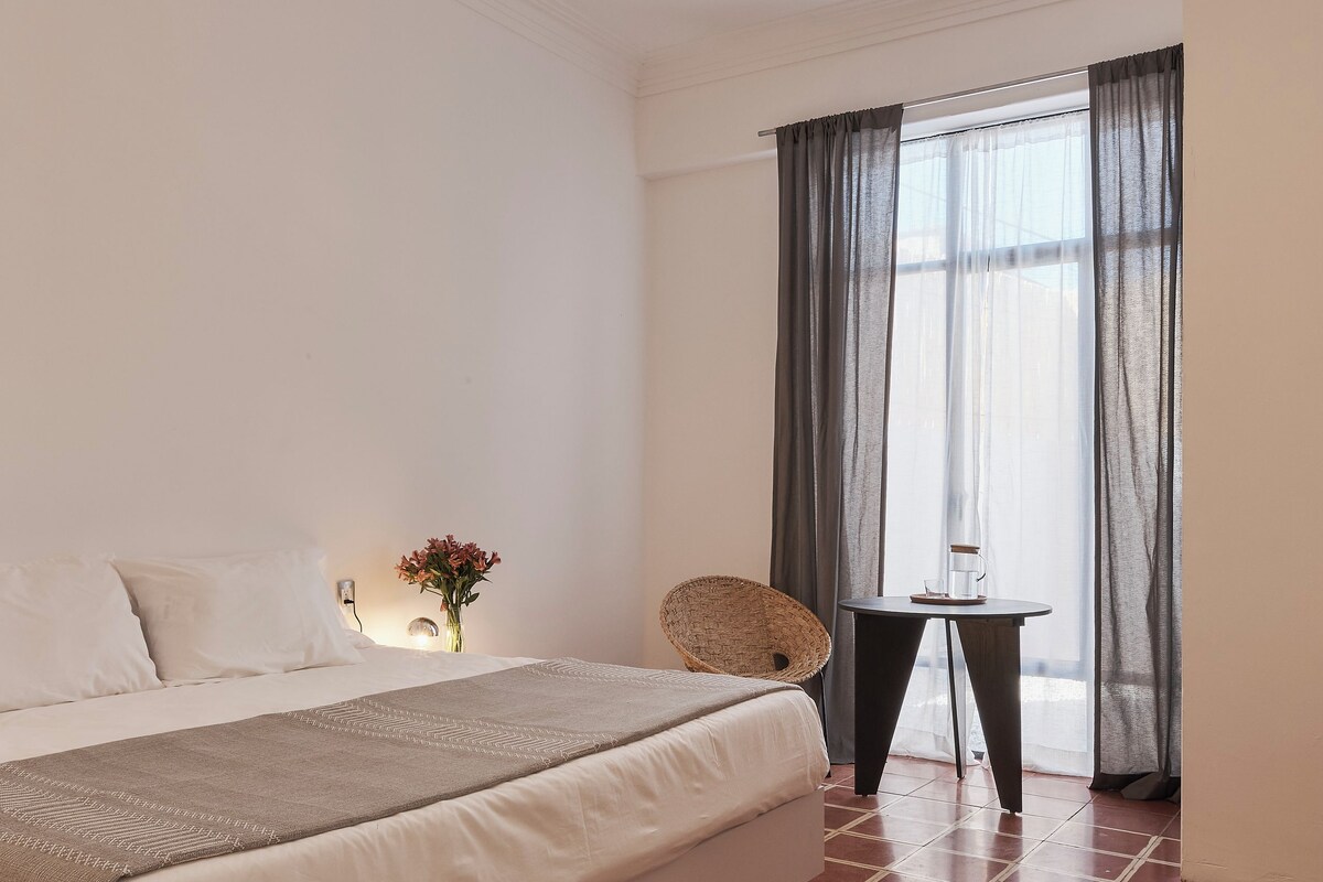 A bedroom with a bed, a ceiling fan, a window with curtains, a small table with a cup, and a vase with flowers.