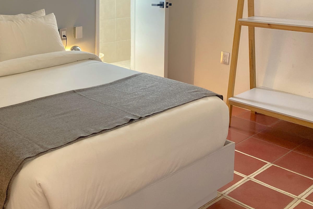 Un lit bien fait, avec du linge de lit blanc et une couverture grise, une table de chevet avec une lampe, et un sol carrelé.