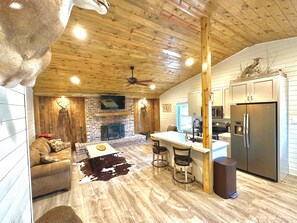 A rustic cabin interior with a fireplace, wooden ceiling, and a kitchen area with a refrigerator and bar stools.