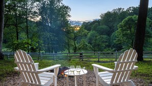 Two Adirondack chairs facing a small table with two glasses of white wine, set on a gravel surface with a fire pit in the background, surrounded by a forested area.
