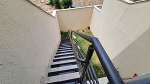 Una escalera estrecha con escalones blancos y negros, flanqueada por paredes color beige y una barandilla de metal.