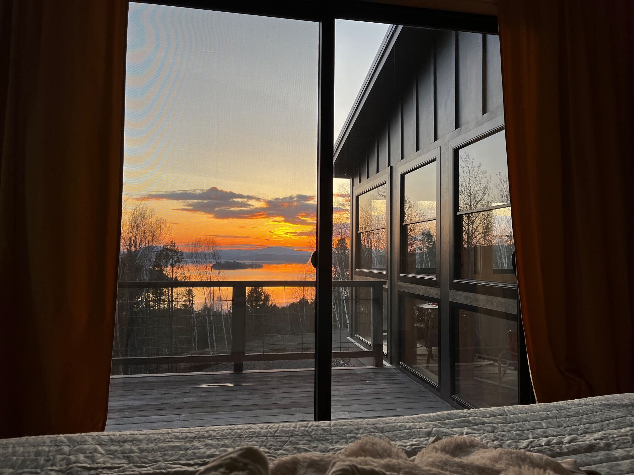 A bedroom with a large window overlooking a deck and a sunset view.