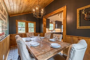 Un comedor con una mesa de madera dispuesta para seis personas, rodeada de sillas tapizadas. Un candelabro cuelga del techo y hay un adorno de cuernos enmarcado en la pared.