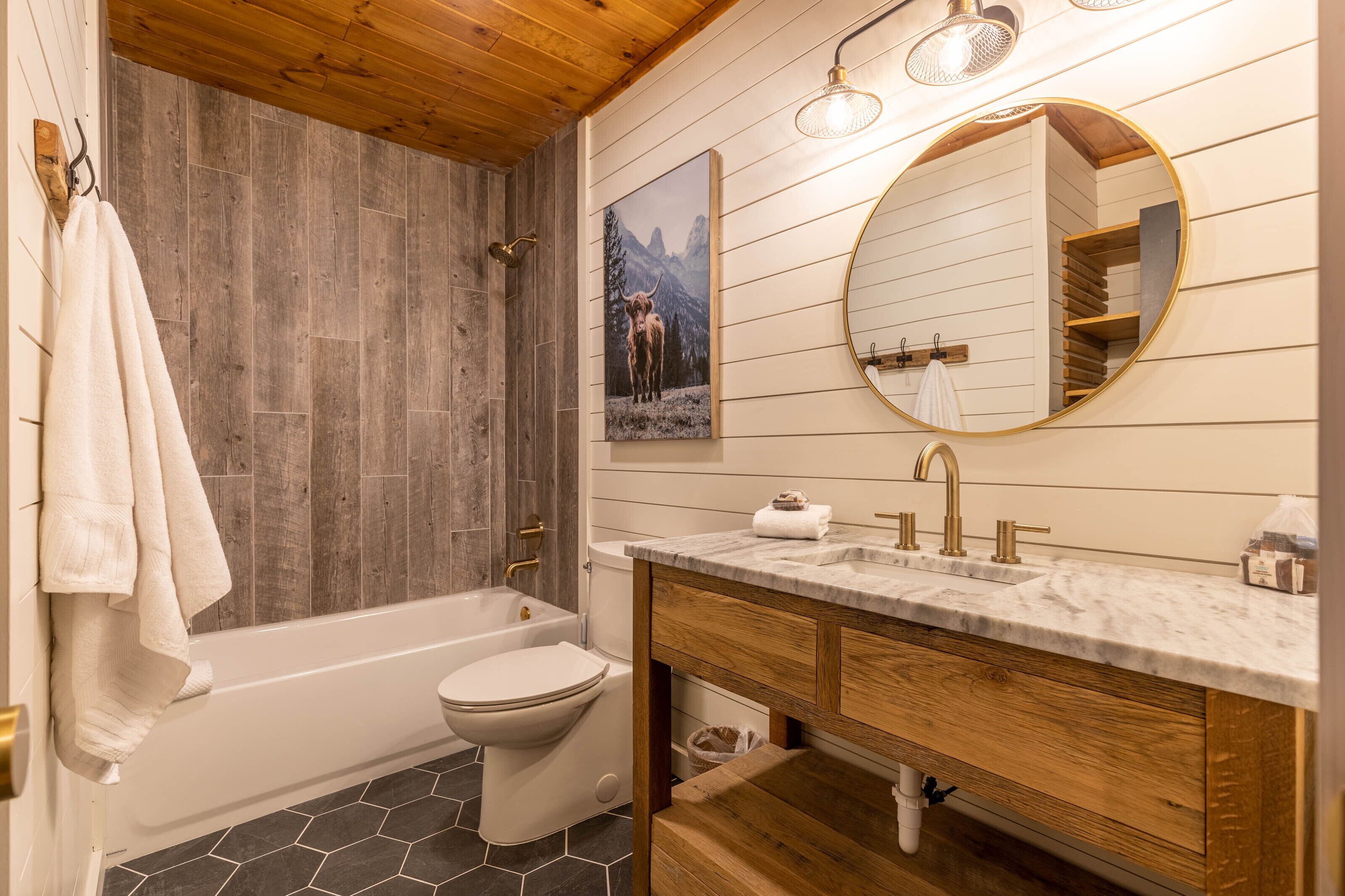 Een badkamer met een houten wand, een bad, een toilet, een wastafel met een marmeren blad en een ronde spiegel.