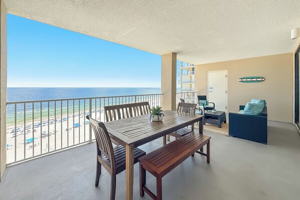 A balcony with a view of the beach, a wooden table with chairs, a bench, and a sofa.